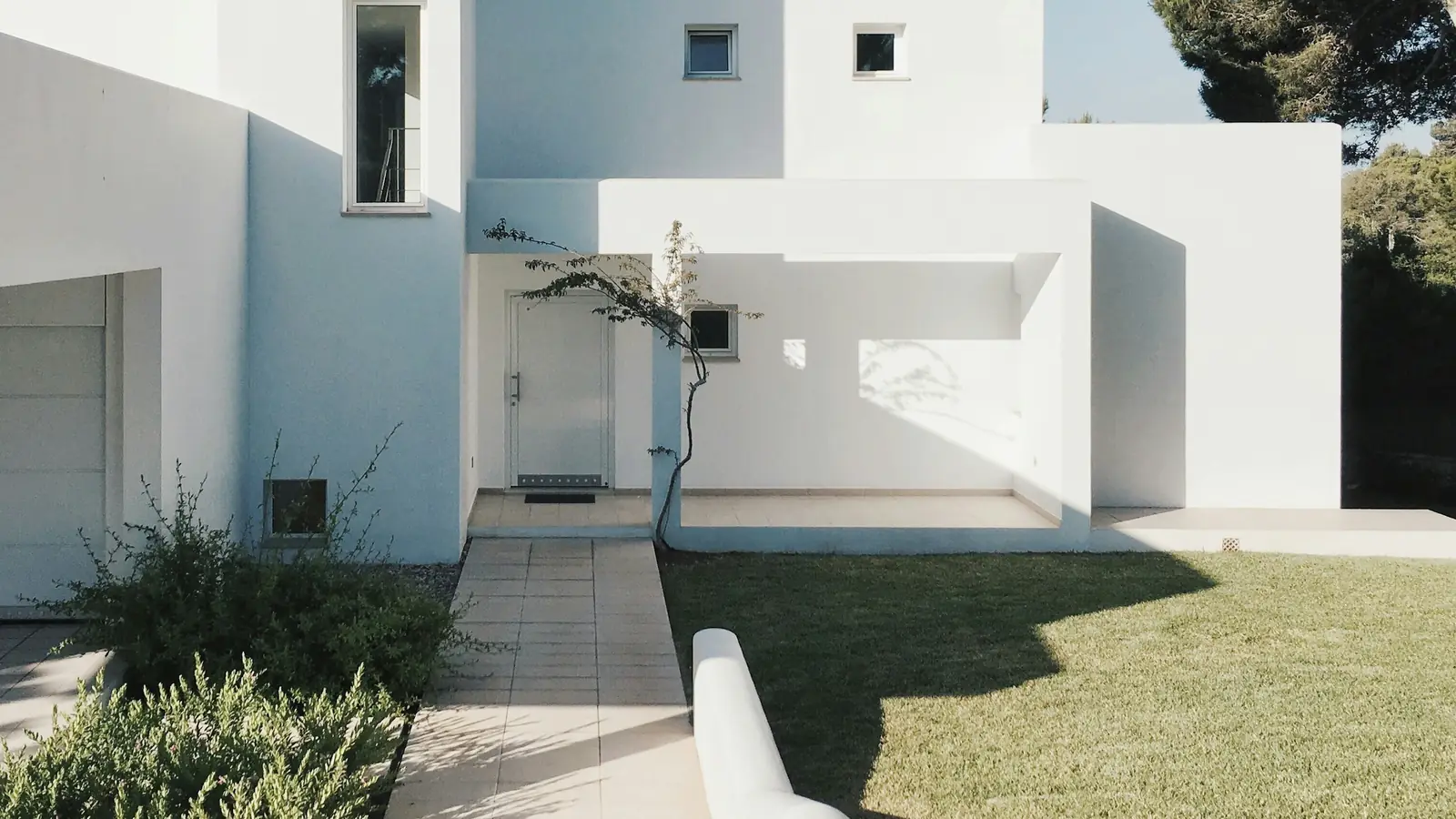 Casa unifamiliar mediterránea de fachada blanca junto a un pino, con jardín y acceso peatonal
