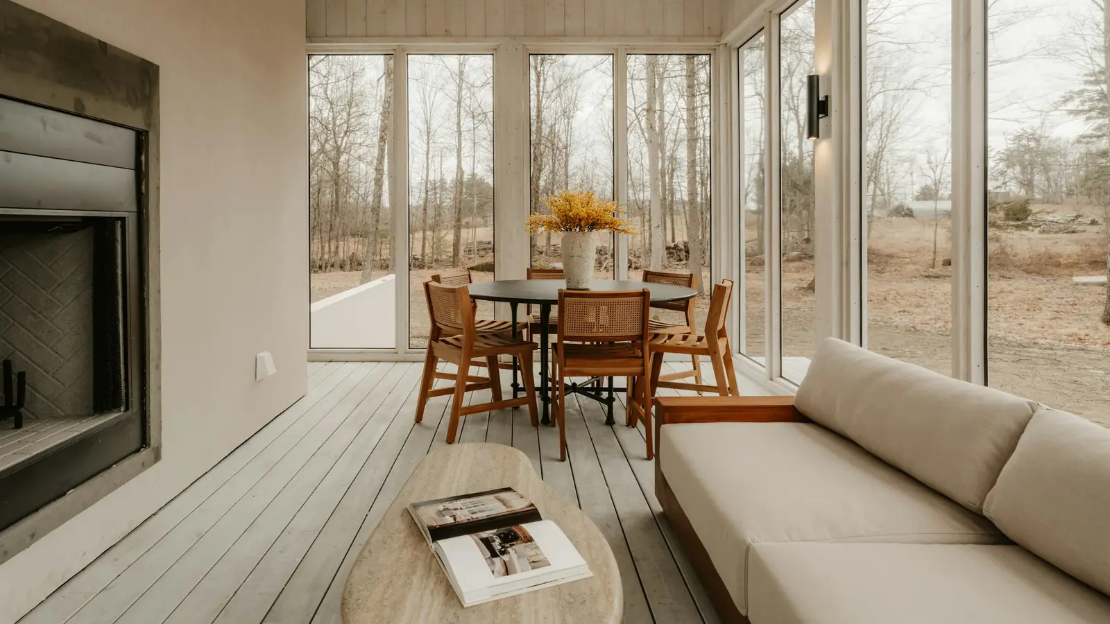 Salón acristalado con chimenea, sofá y comedor de madera rodeado de árboles